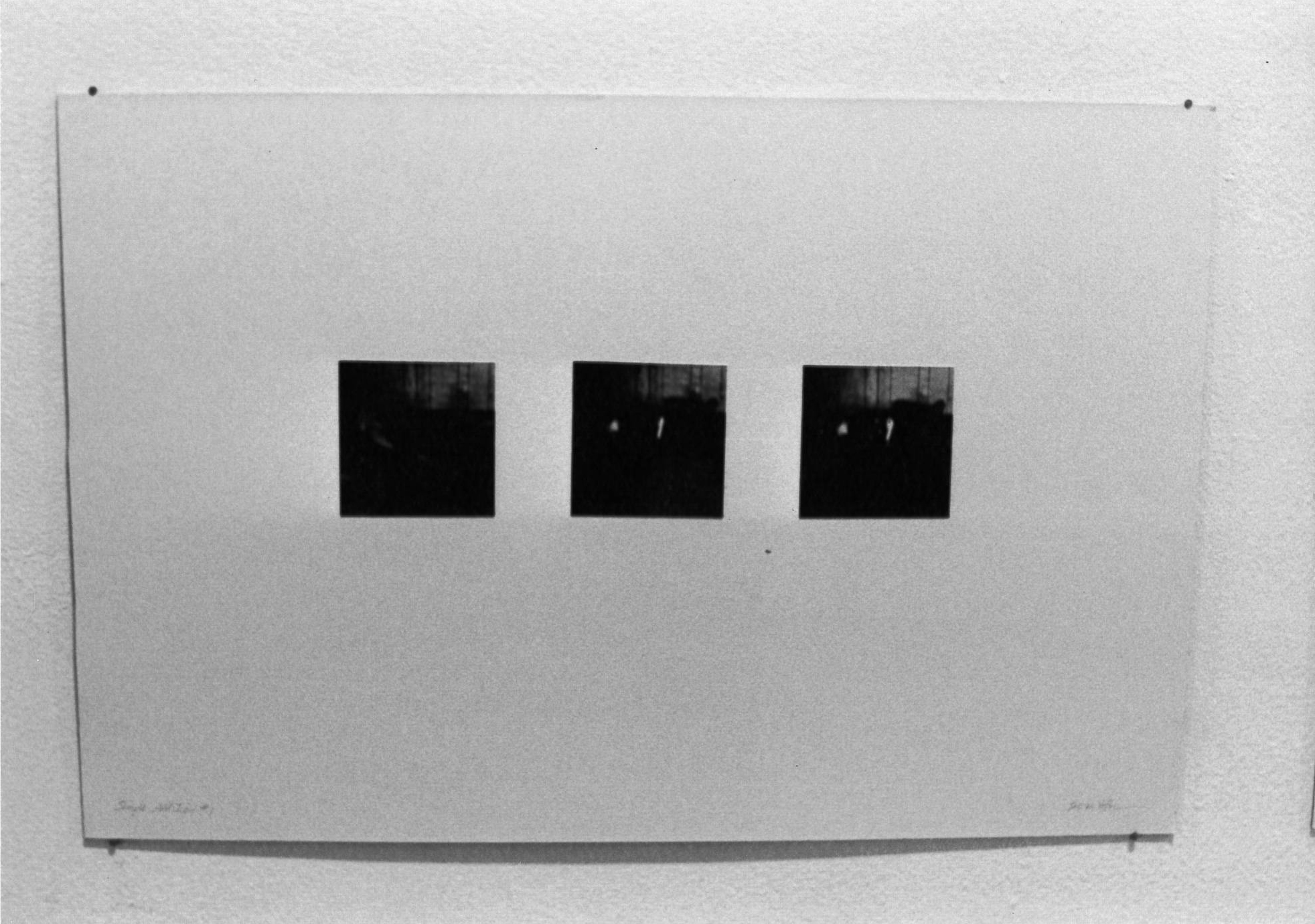 A black and white installation view of three square photographs with wide white matting attached to a wall.
