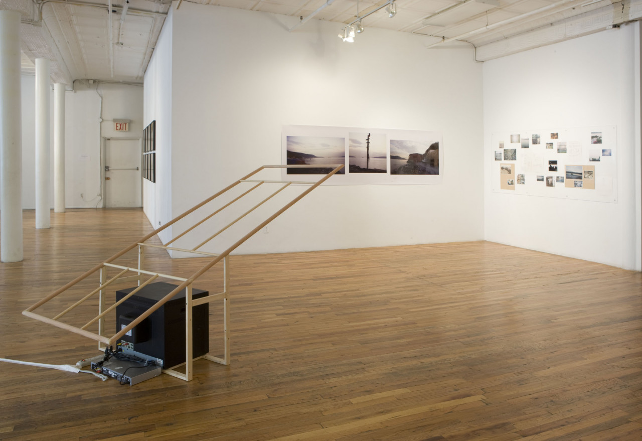 A television sits on the floor, encaged by a wooden structure extending at an angle into the air. On the walls opposite, there are assorted photographs of varying sizes, showing primarily landscapes.