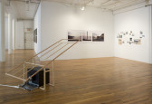 A television sits on the floor, encaged by a wooden structure extending at an angle into the air. On the walls opposite, there are assorted photographs of varying sizes, showing primarily landscapes.