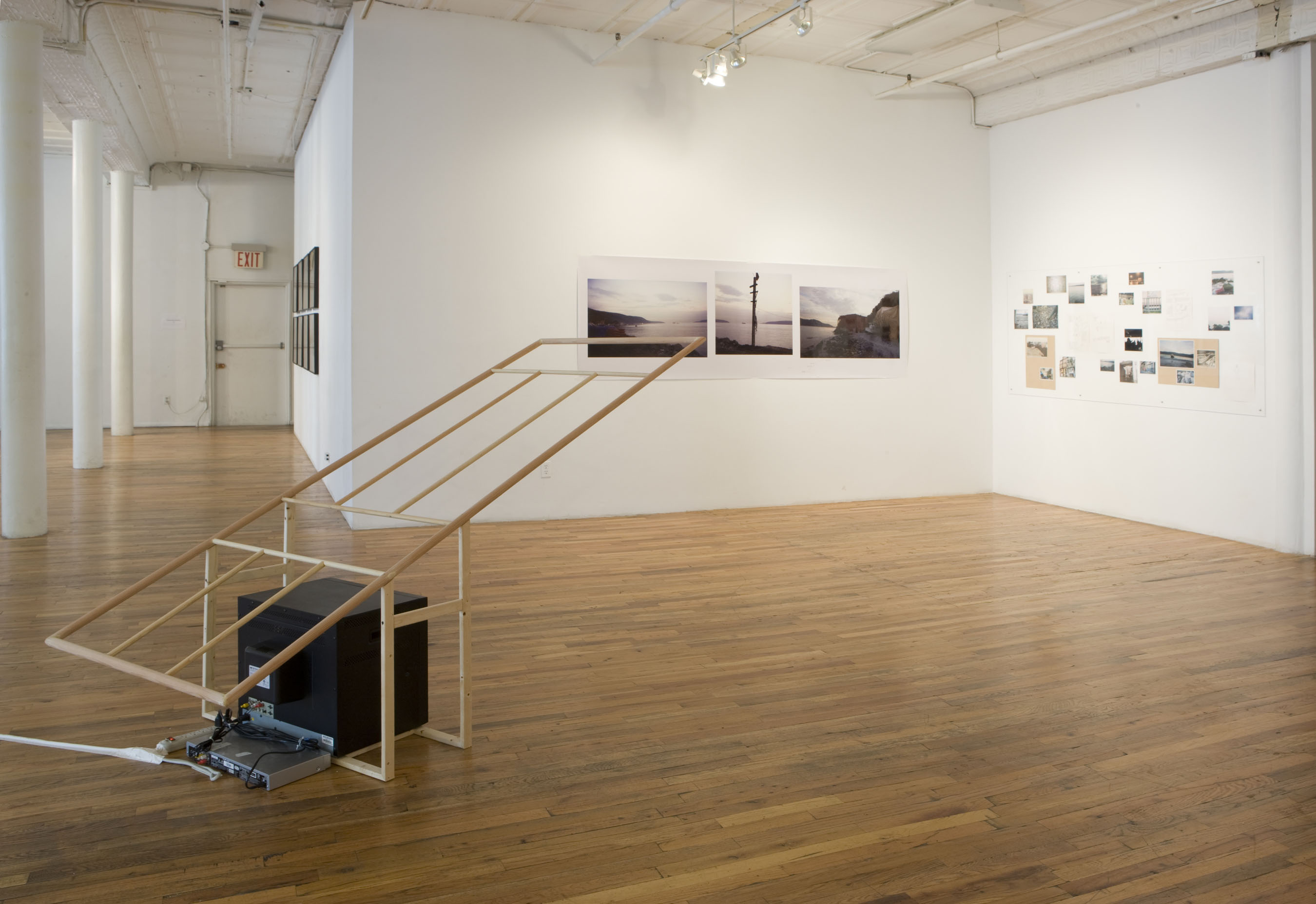 A television sits on the floor, encaged by a wooden structure extending at an angle into the air. On the walls opposite, there are assorted photographs of varying sizes, showing primarily landscapes.