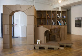 An archway made of cardboard boxes connected to a bookcase and curved desk, also made of cardboard. A white lucky waving cat is placed at the bottom left side of the desk. The desk and bookcase are supported by a platform, underneath of which a pair of slippers are visible.