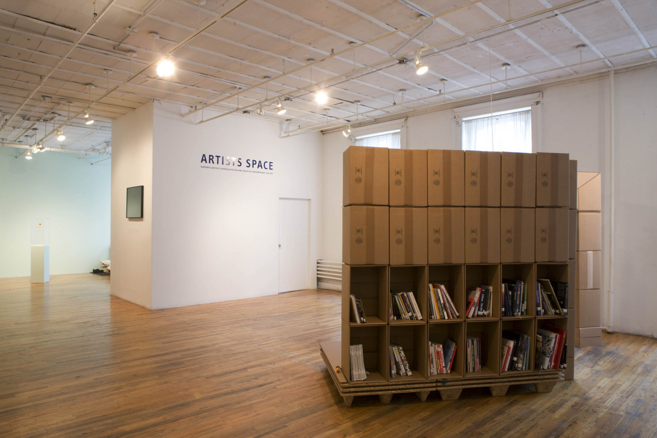 A bookshelf constructed from cardboard boxes sits slightly raised off the ground on a low platform. The top two rows face away from the camera, while the bottom two face out, filled with books. Opposite that is a white wall with blue text reading "Artists Space."