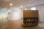 A bookshelf constructed from cardboard boxes sits slightly raised off the ground on a low platform. The top two rows face away from the camera, while the bottom two face out, filled with books. Opposite that is a white wall with blue text reading 