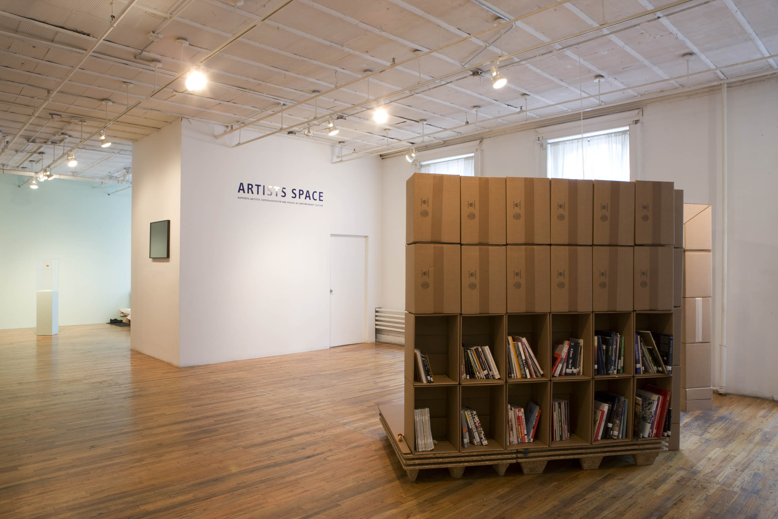 A bookshelf constructed from cardboard boxes sits slightly raised off the ground on a low platform. The top two rows face away from the camera, while the bottom two face out, filled with books. Opposite that is a white wall with blue text reading 