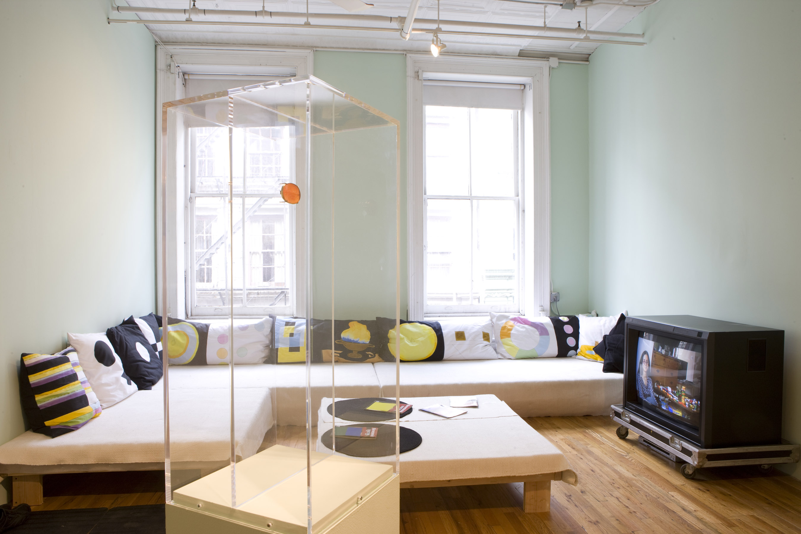 A low futon couch on a wooden platform with multicolored pillows is fit into the corner of a room with two windows. There is a short table with two placemats and brochures, and a television. In the foreground, there is a white plinth with a glass top housing an orange object.