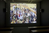 A dark room with a video projection on the wall and several benches. The image shows a crowd of people, some children and some adults, sitting on grass and watching a guitar player.