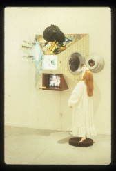 A sculpture of a woman stands on a pedestal looking into a glass window pyrimidal construction with a large bug on top and a TV monitor underneath.