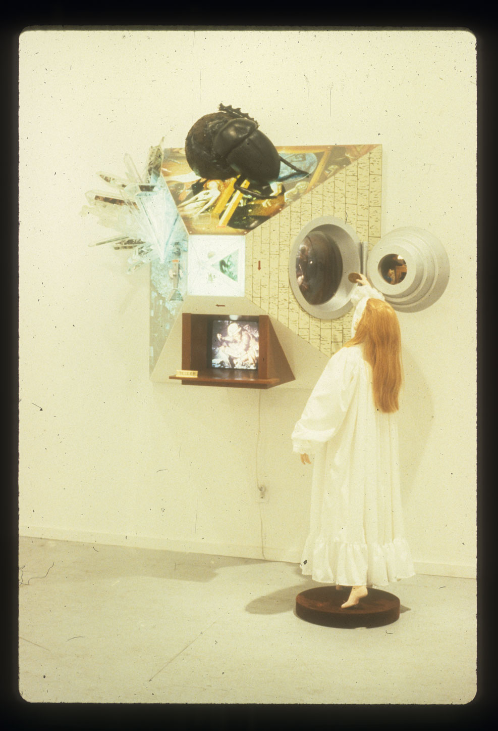 A sculpture of a woman stands on a pedestal looking into a glass window pyrimidal construction with a large bug on top and a TV monitor underneath.