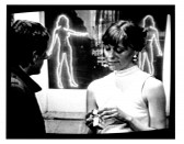 A woman looks down and opens a package in her hands while a man looks at her. Peep show neon signs can be viewed in the background.