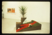 A piece of landscaping appears on the gallery floor with flowers and plants coming up from it in a stair-like fashion. A painting appears in the background in a white-walled gallery space.