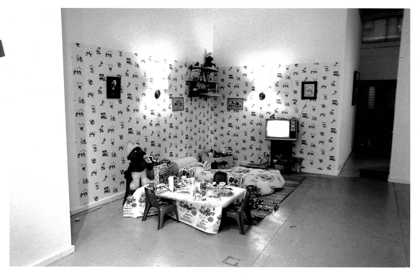 A table with household items like cereal and plates and cups appear on it; next to this lies a bed and a television set. Wall paper is visible along with small framed pictures on the wall in a gallery setting.