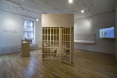 A freestanding room, constructed from wood and resembling a prison cell, sits in the center of a gallery space, its door ajar. A model of a building sits on a pedestal to the left of the wooden prison cell. On the wall, a vinyl quote reads: WHAT KIND OF HOUSE DOES / A MAN WHO HAS LIVED IN A / SIX-FOOT BY NINE-FOOT CELL / FOR OVER 35 YEARS DREAM OF?