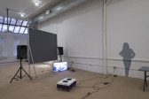 To the right, a gray silhouette of a woman with her hands on her hips is installed on a white wall. Exposed wires run from an electrical socket to the left of her legs to a box on the ground. A television monitor sits on the ground in front of the back of a projection screen positioned between two standing speakers.