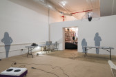 Gray silhouettes are displayed on adjacent walls. Miscellaneous items are displayed on gray tables. A microphone and its extended wire lays on the floor in front of a speaker.