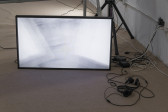 A television monitor positioned on the ground with its wiring exposed. A pair of headphones rests on the ground to the right of the monitor. It displayes a still image of a washed-out portrait that implies movement through the blurring and repeititon of the face.