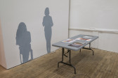 Two gray silhouettes displayed next to each other on a white wall. In front of them, a gray table displays colorful documents.