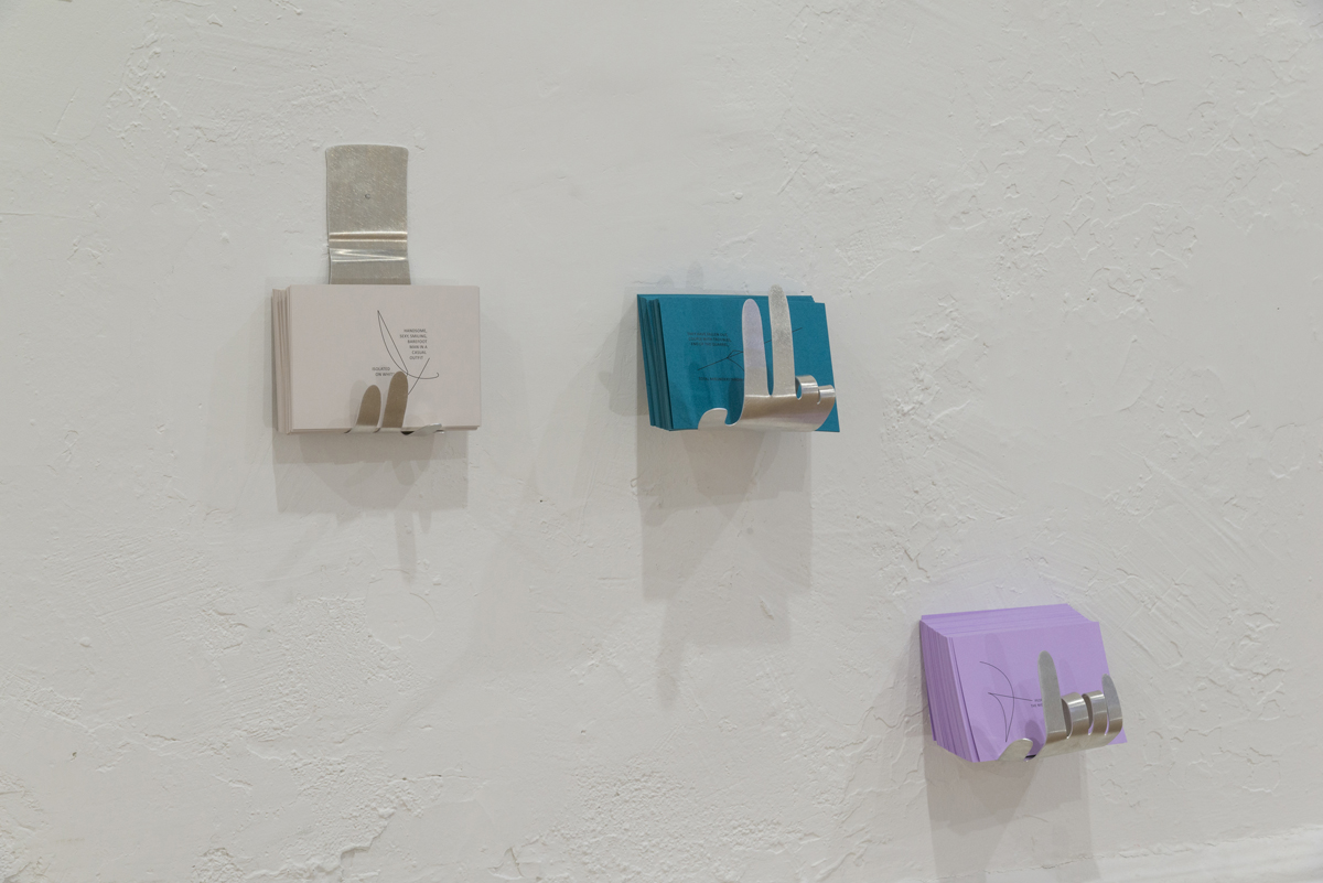 Three metal cutouts of hands, each displaying a different finger expression, are installed on a white wall. Each hand holds stacks of white, blue, and purple cards.