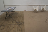 The floor of a gallery space divided into two sections, with wooden flooring on the left and carpeted flooring on the right. On the left, a fold-out table displays colorful documents. On the right, four minature copper squares sit on the ground next to two cream-colored foldable chairs. Multiple wires extend across the floor and under the carpet.