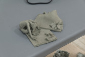 A miniature clay sculpture sitting on a gray table. The sculpture depicts a woman folding over, cross-legged, on carpet. Miscelaneous items are dispersed across the carpets.