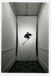 A photograph of a hallway with a white piece of cloth hanging from the ceiling to the floor. Printed on the cloth is a figure dressed in black and bent at the waist holding a long stick.