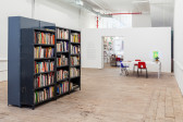 An angled view of grey, metal book shelves sat towards left frame in the center of a room. The shelves are populated with books of various sizes and colors. In the background, right frame, a table with charis and books sits infront of a wall opening leading to an entrance/exit.