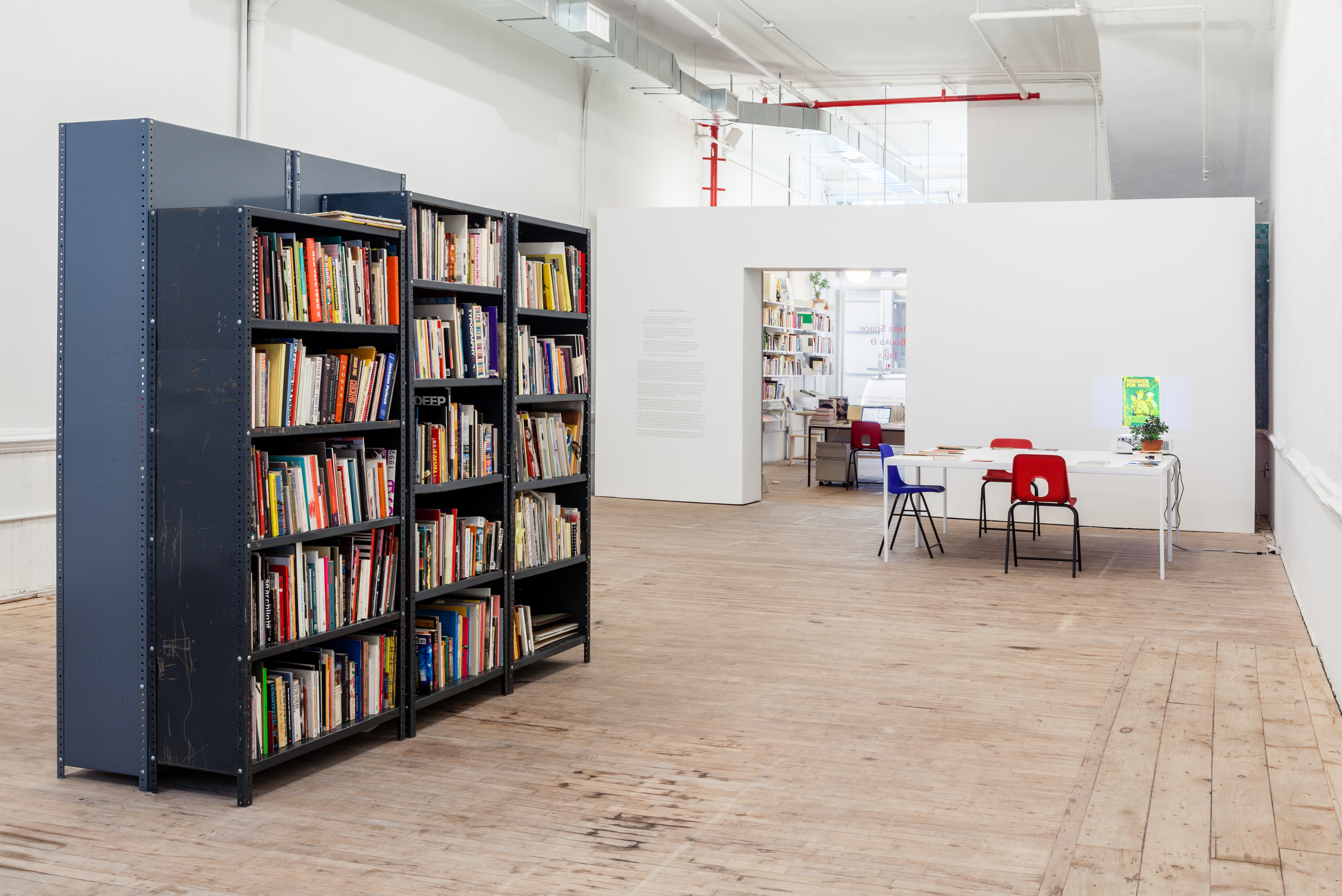 An angled view of grey, metal book shelves sat towards left frame in the center of a room. The shelves are populated with books of various sizes and colors. In the background, right frame, a table with charis and books sits infront of a wall opening leading to an entrance/exit.