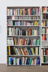 A seven shelf, white, book case is housed full of various colorful and sized books.