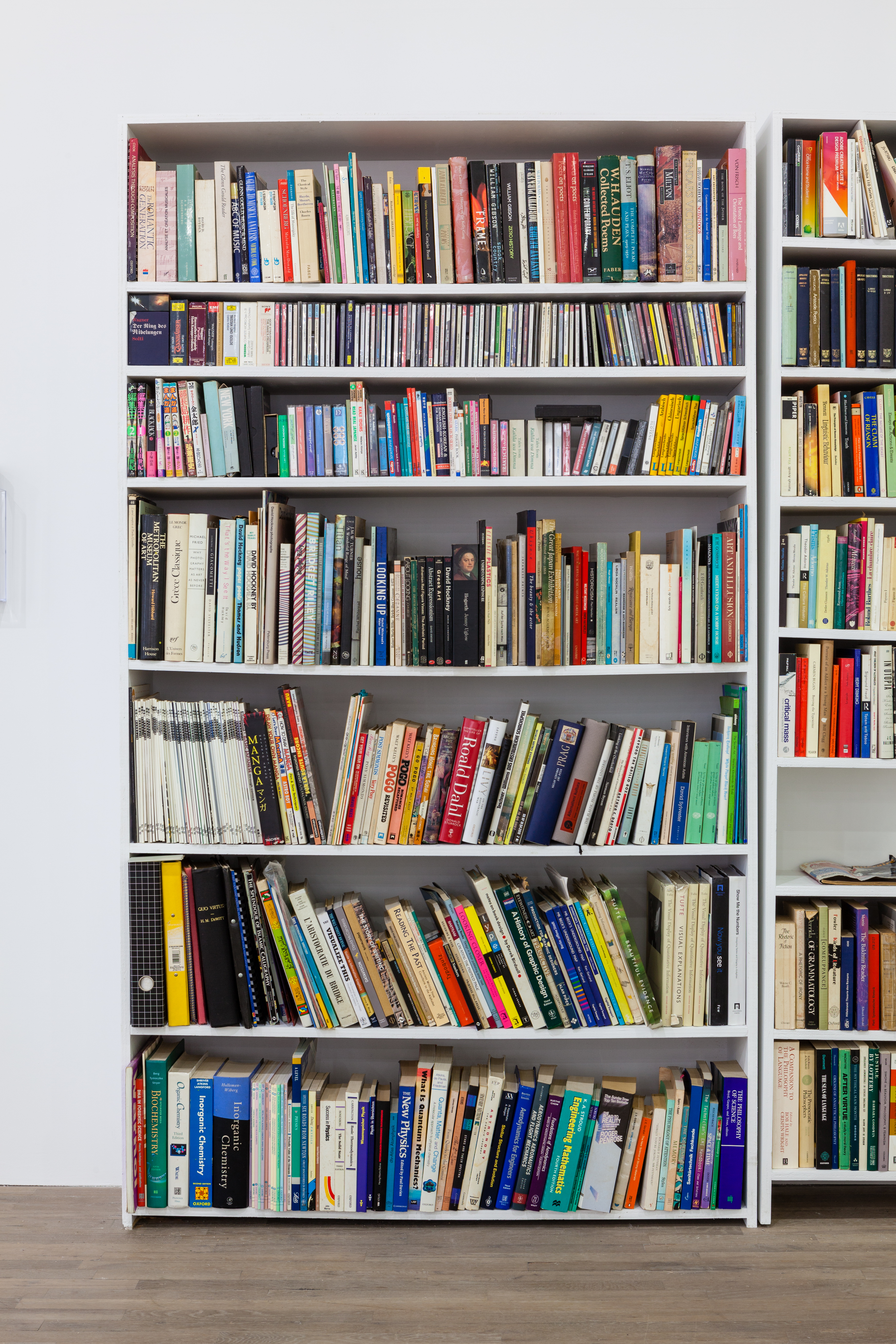 A seven shelf, white, book case is housed full of various colorful and sized books.