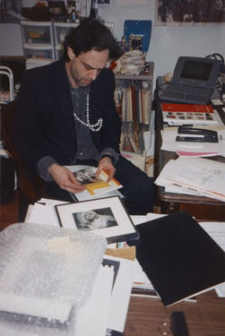 Colin de Land sat at a desk, looking at paper materials in his lap. Various objects, documents, and books sit opened around him.