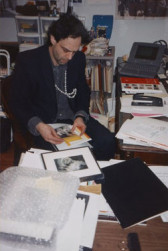 Colin de Land sat at a desk, looking at paper materials in his lap. Various objects, documents, and books sit opened around him.