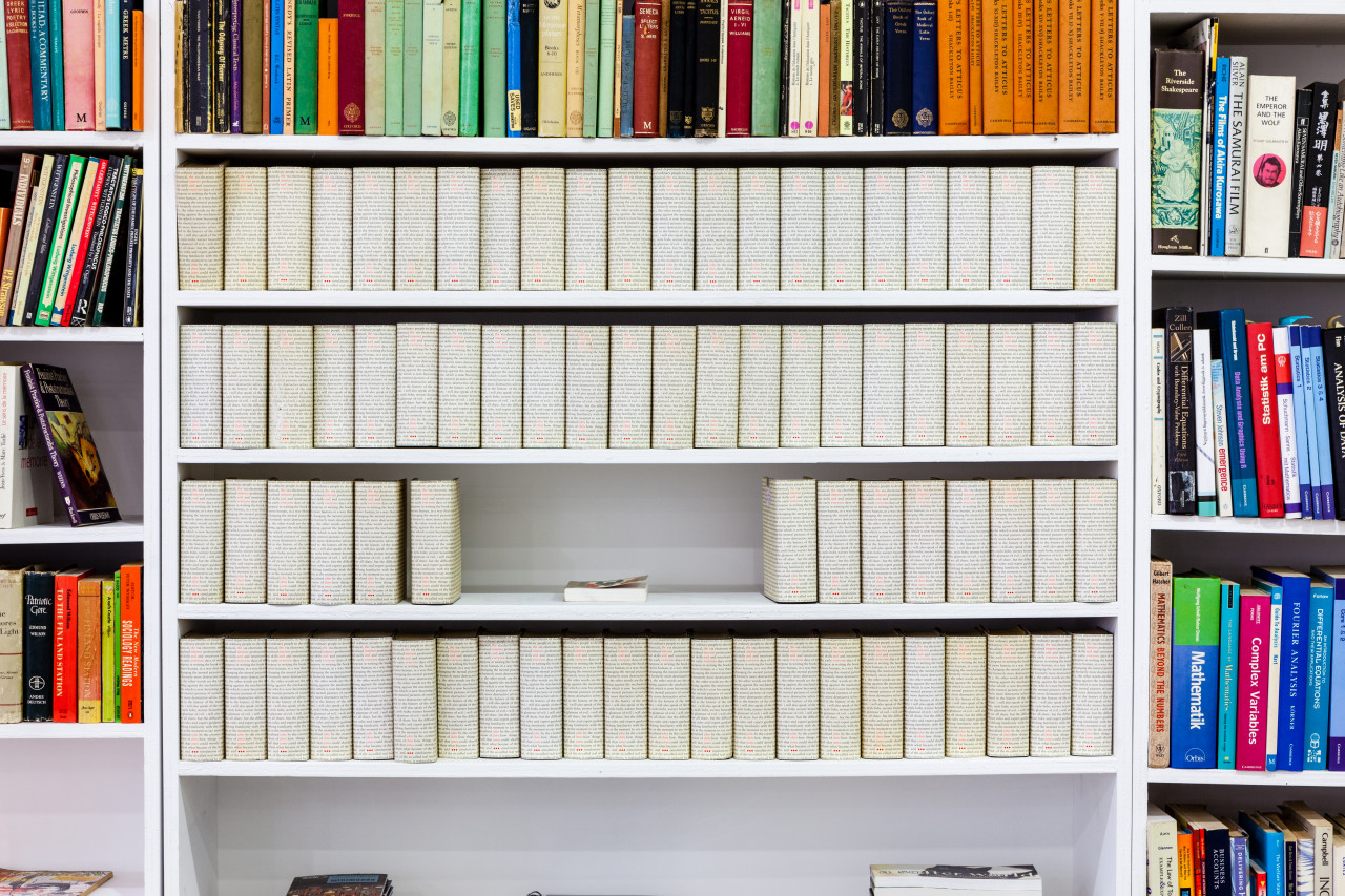 A book shelf, of the center are four shelves, filled with books whose covers contain small printed text. In the center of the second from the bottom shelf, a book sits horizontal.