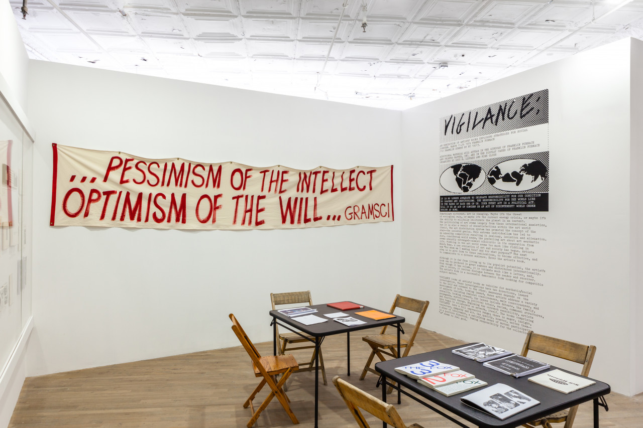 An angled view of a room with a set of two tables and chairs, books resting on the tables, in the center. On the back wall hangs a banner in red painted text, reads "...Pessimism of the intellect, Optimism of the will... Gramsci." On the right adjacent wall, text covers the whole height, the title reading, "Vigilance:"