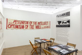 An angled view of a room with a set of two tables and chairs, books resting on the tables, in the center. On the back wall hangs a banner in red painted text, reads 