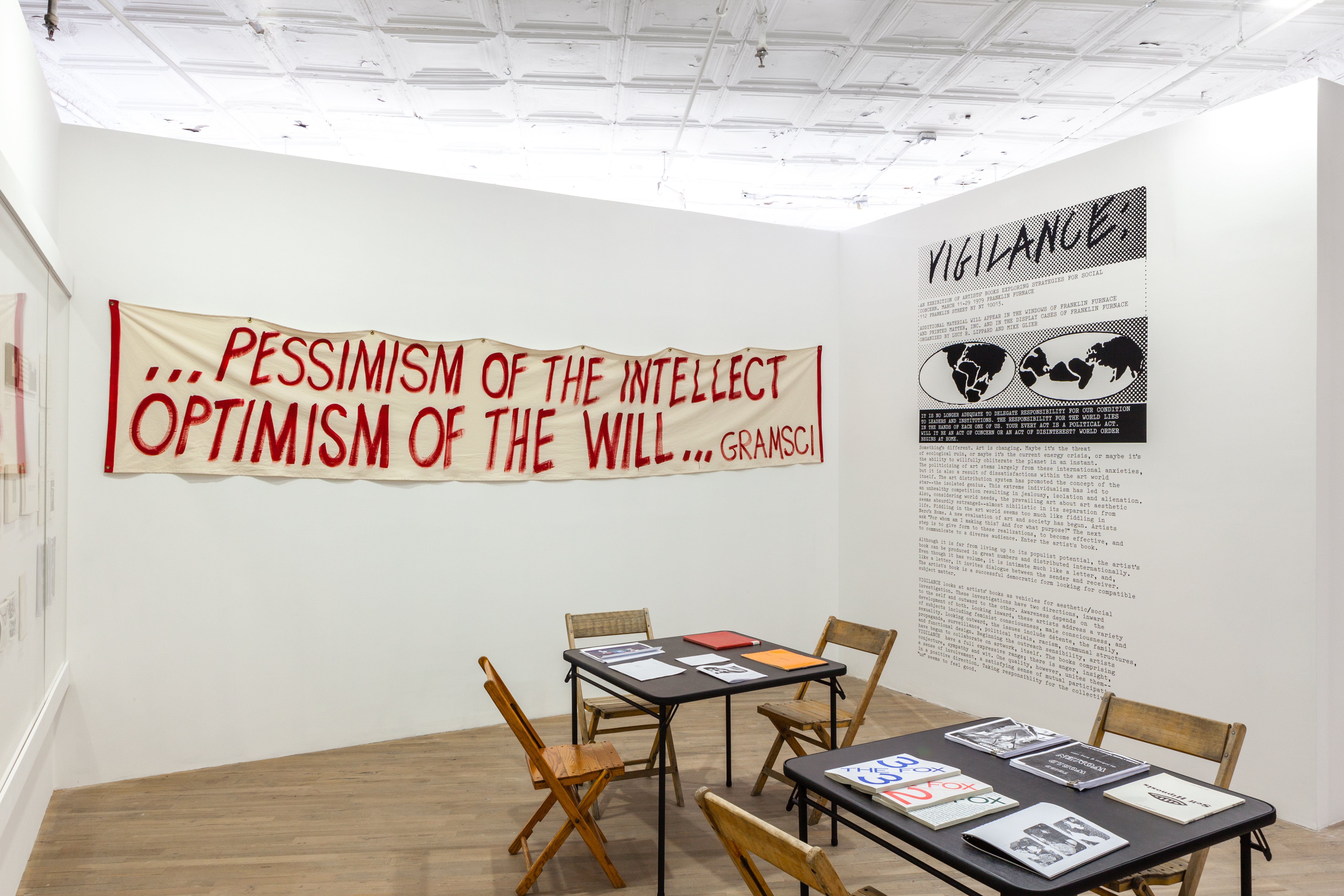 An angled view of a room with a set of two tables and chairs, books resting on the tables, in the center. On the back wall hangs a banner in red painted text, reads 