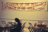 A woman sits, left of center, at a table filled with books. On the wall behind her, a banner with red text that reads, 