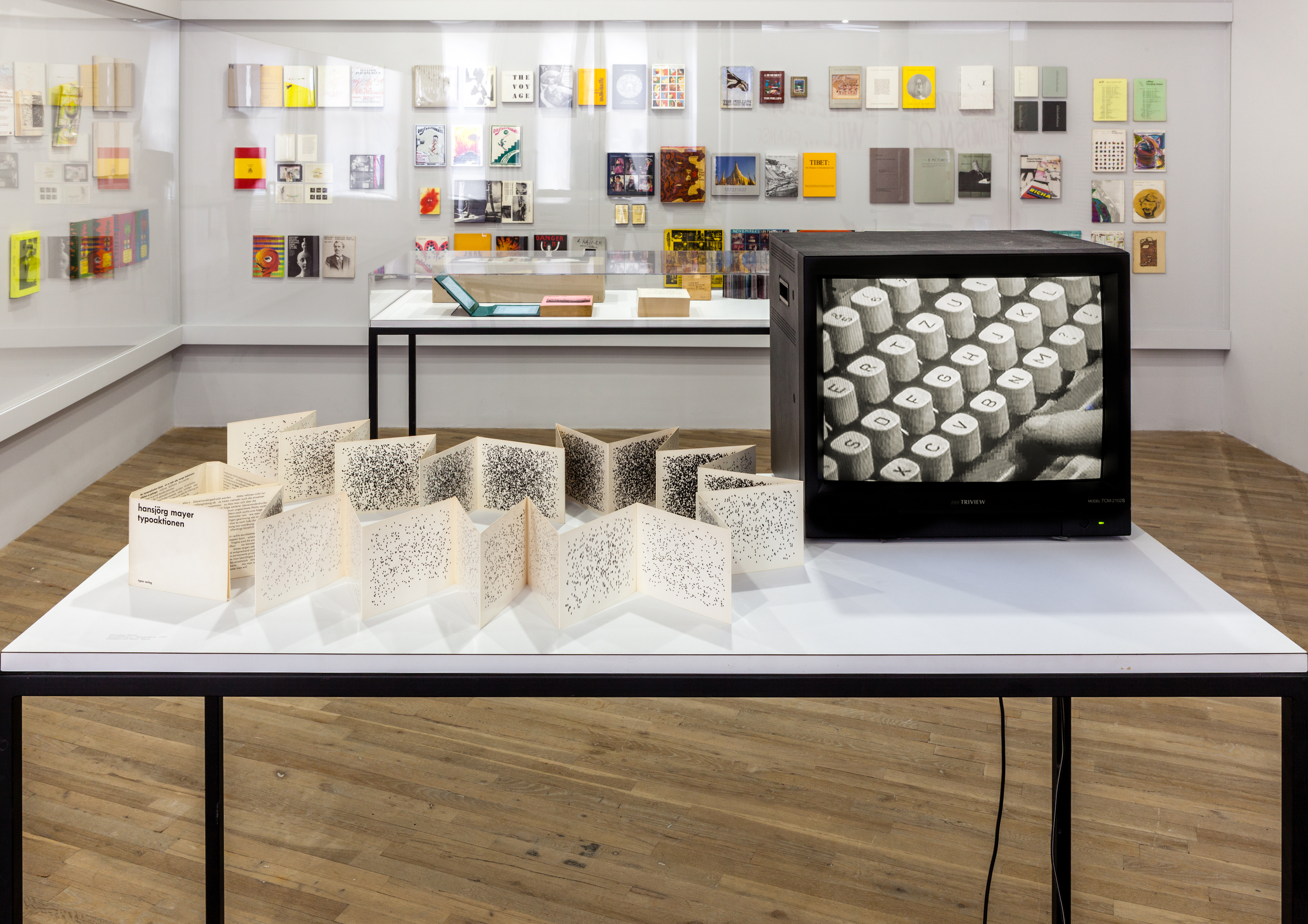 On a white table, to the left, sits a flutter book with a dot illustration increasing in density through the pages. To the right, a TV monitor displays a black and white image of a finger pressing the space bar on a type writer.