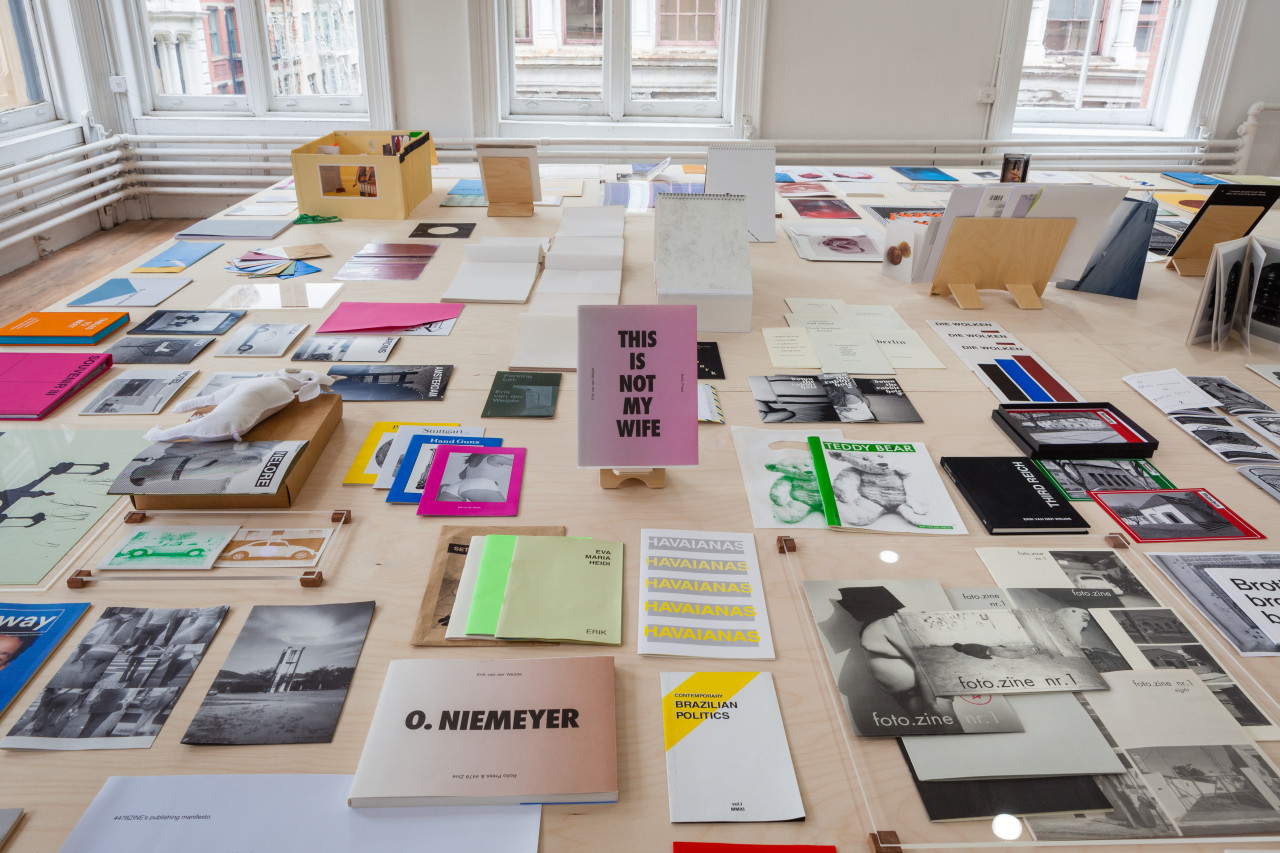 An angled view of a wooden table containing various papers, books, and documents displaying text and images. Windows to the street surround the background.