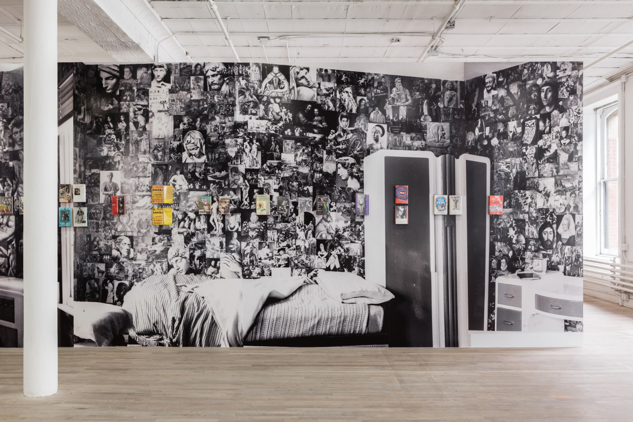 A series of books sit at eye level on a wall that displays a black and white photographic collage graphic behind them.