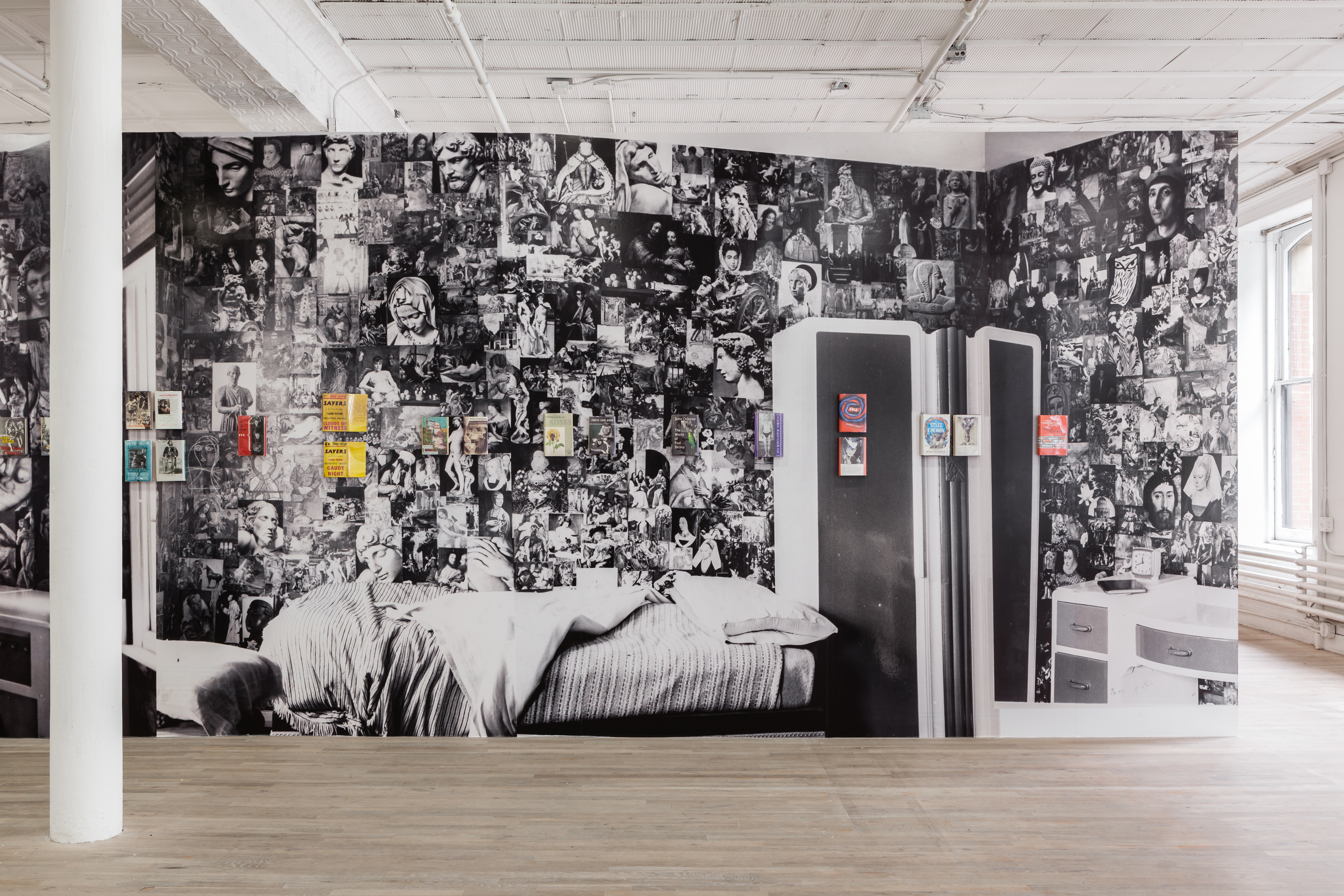 A series of books sit at eye level on a wall that displays a black and white photographic collage graphic behind them.