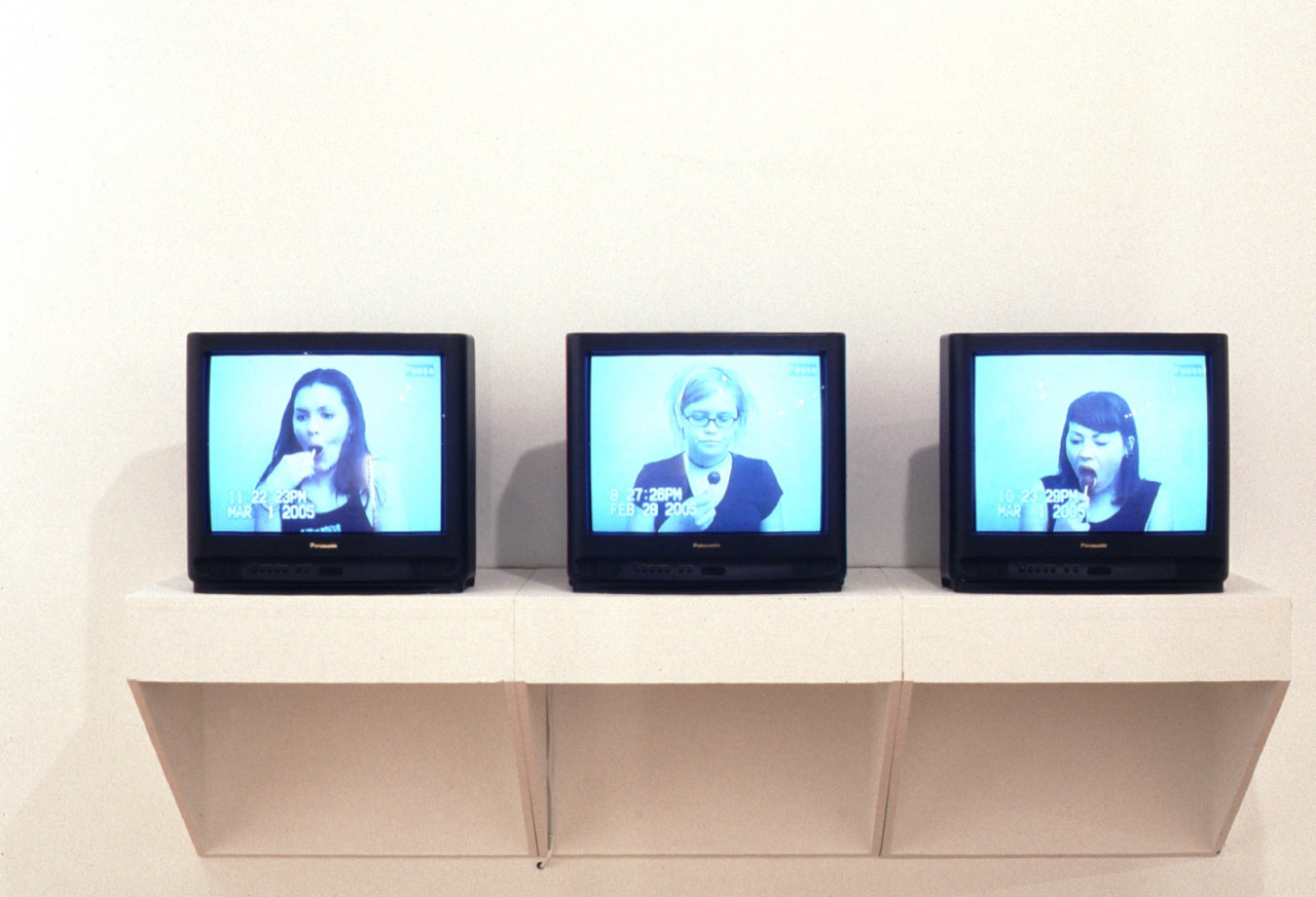 Three small televisions sit on a white shelf. Each displays a timestamped video of a person eating a lollipop.