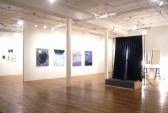 A gallery space with two columns. On the right side of the columns is a small platform from which two black curtains hang. Behind that are two stools underneath a mirror mounted on the wall. On the other side of the columns, four paintings hang on a white wall in a line. Just visible beyond the end of the wall is another space with additional paintings hung on that wall.