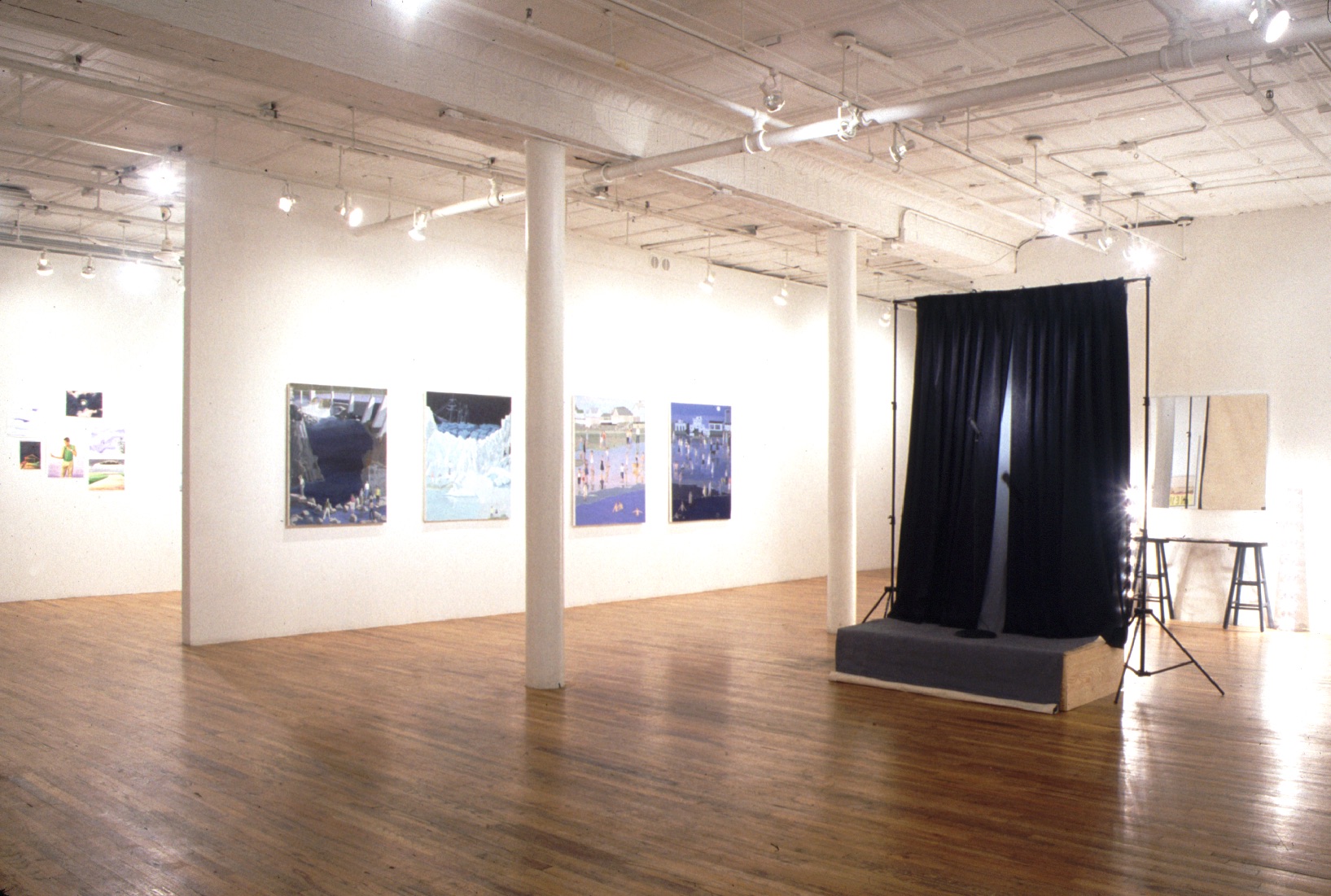 A gallery space with two columns. On the right side of the columns is a small platform from which two black curtains hang. Behind that are two stools underneath a mirror mounted on the wall. On the other side of the columns, four paintings hang on a white wall in a line. Just visible beyond the end of the wall is another space with additional paintings hung on that wall.