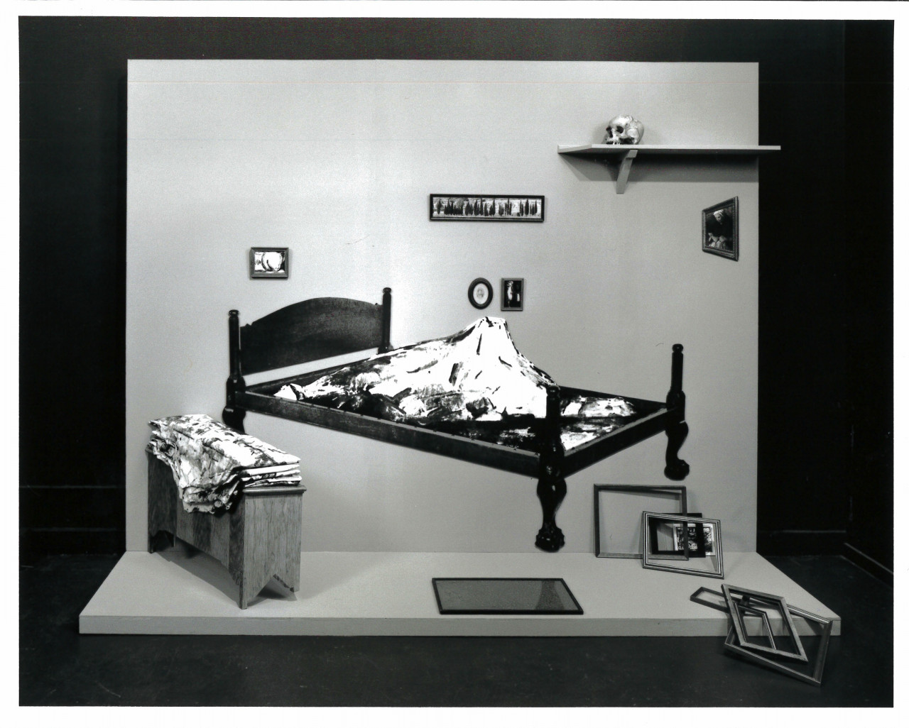 A bed with a mountain at its center in a partially constructed room with a floor and a single wall. Surrounding it are several framed paintings and empty frames, a bench with fabric piled on it, and a high shelf with a skull.