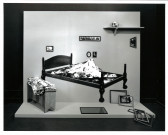 A bed with a mountain at its center in a partially constructed room with a floor and a single wall. Surrounding it are several framed paintings and empty frames, a bench with fabric piled on it, and a high shelf with a skull.