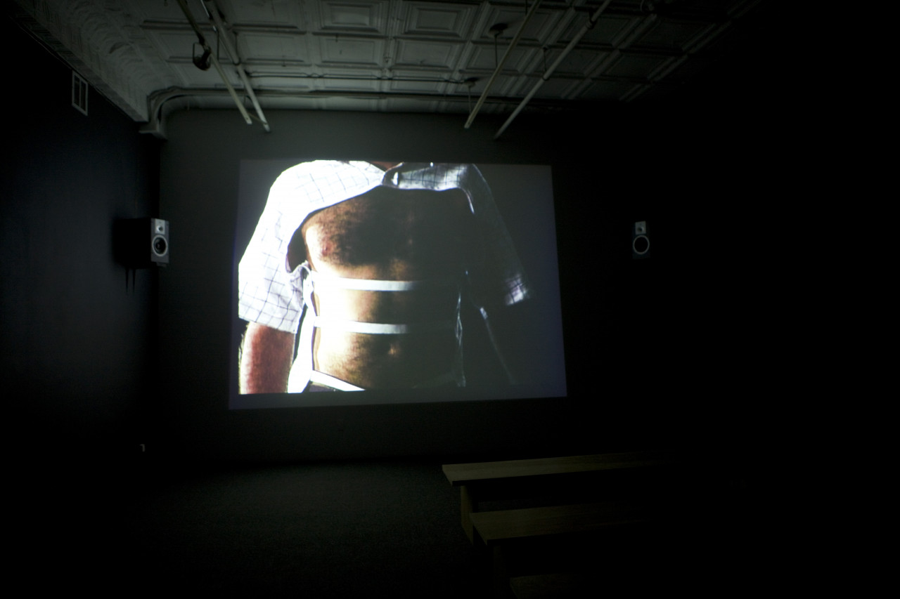 A dark room with a screen projection showing an image of a man with a partially cut out button down shirt. The sleeves and top button are intact, with the chest and torso bare except for three elastic strips spaced out spanning the torso.