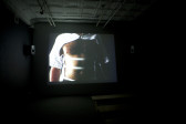 A dark room with a screen projection showing an image of a man with a partially cut out button down shirt. The sleeves and top button are intact, with the chest and torso bare except for three elastic strips spaced out spanning the torso.
