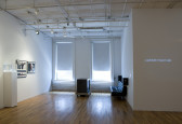 A room with white walls, with two windows on the back wall with shades pulled halfway down. The wall to the left of the windows has three framed photographs, and a white plinth with a glass top. In front of the windows, a television on a low table faces sideways toward a low black couch. On the far right side, a protruding corner of a wall has white text projected on it reading 