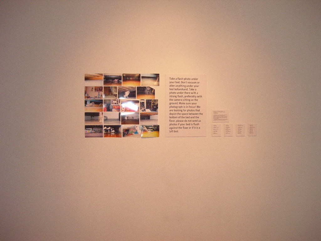 20 photographs arranged in 5 rows of 4 on a white wall. The photos show various shots taken under beds. There is descriptive wall text to the right of the photos.