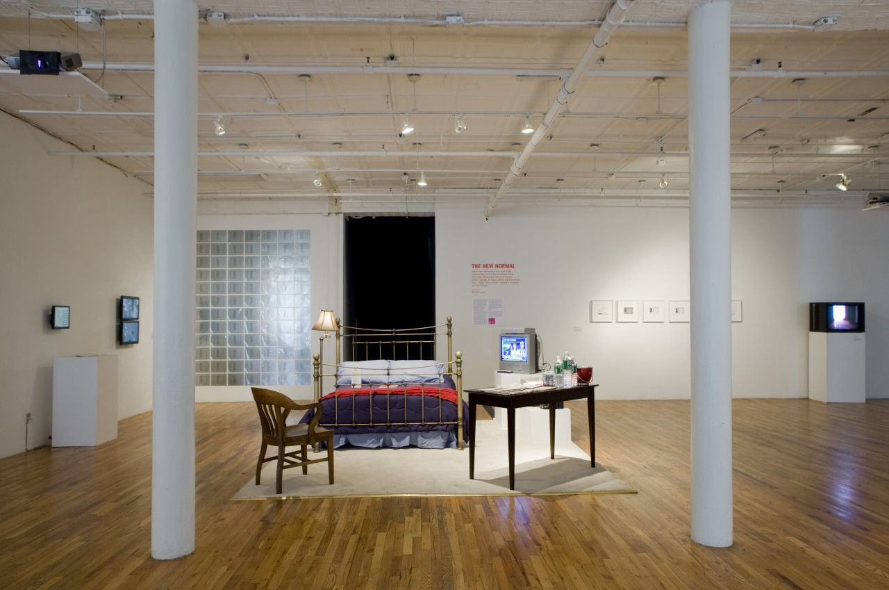 A bedroom scene set up in the middle of an open room, seen between two white poles. There is a beige rug, with a chrome lamp and matching bed frame. The bed has blue sheets with a dark blue comforter cover and a red blanket. There is a wooden chair facing in, and a brown table with assorted bottles on top. On the far side of the bed is a coffee machine, microwave, and television. In the background, various framed images are hung on white gallery walls.