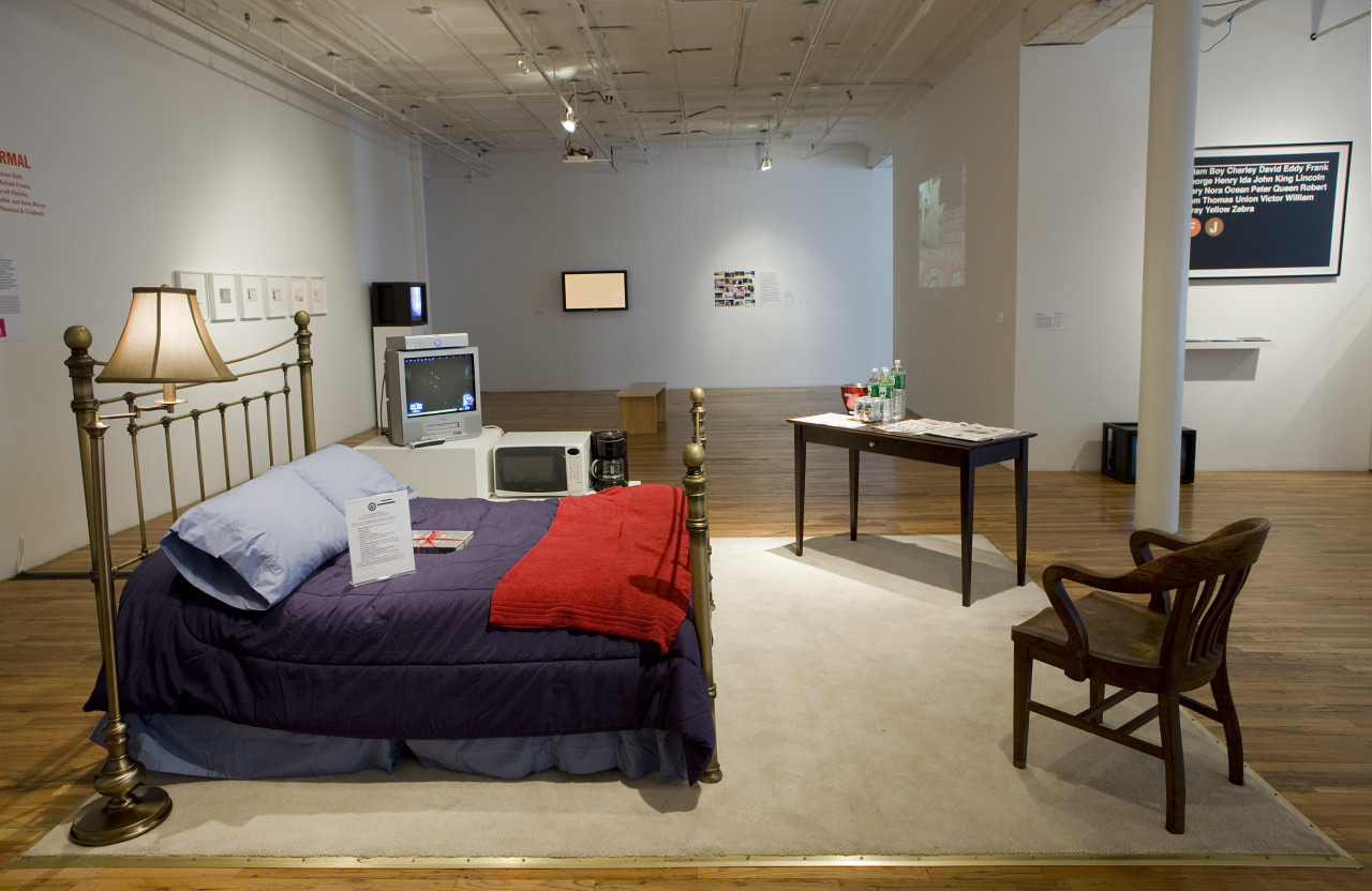 A bedroom scene set up in the middle of an open room. There is a beige rug, with a chrome lamp and matching bed frame. The bed has blue sheets with a dark blue comforter cover and a red blanket. There is a wooden chair facing in, and a brown table with assorted bottles on top. On the far side of the bed is a coffee machine, microwave, and television. In the background, various framed images are hung on white gallery walls.